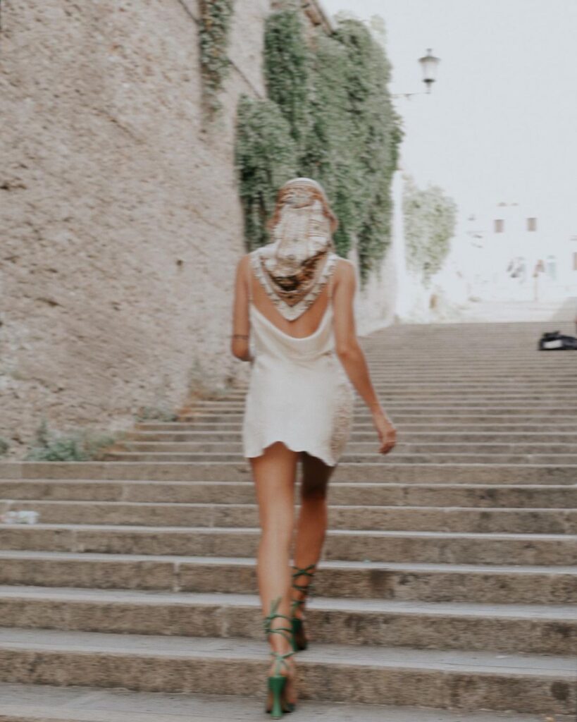 A woman ascends an old stone staircase, her back to the camera, in a timeless Mediterranean setting. She wears a flowing white dress, paired with striking green lace-up heels. A patterned headscarf, artfully wrapped around her hair, adds an air of mystery. The scene is serene, framed by weathered walls and cascading greenery, evoking the rich history and romantic charm of a quaint coastal town. Her movement suggests a journey, an ascent not just of steps, but perhaps towards a new experience or discovery.