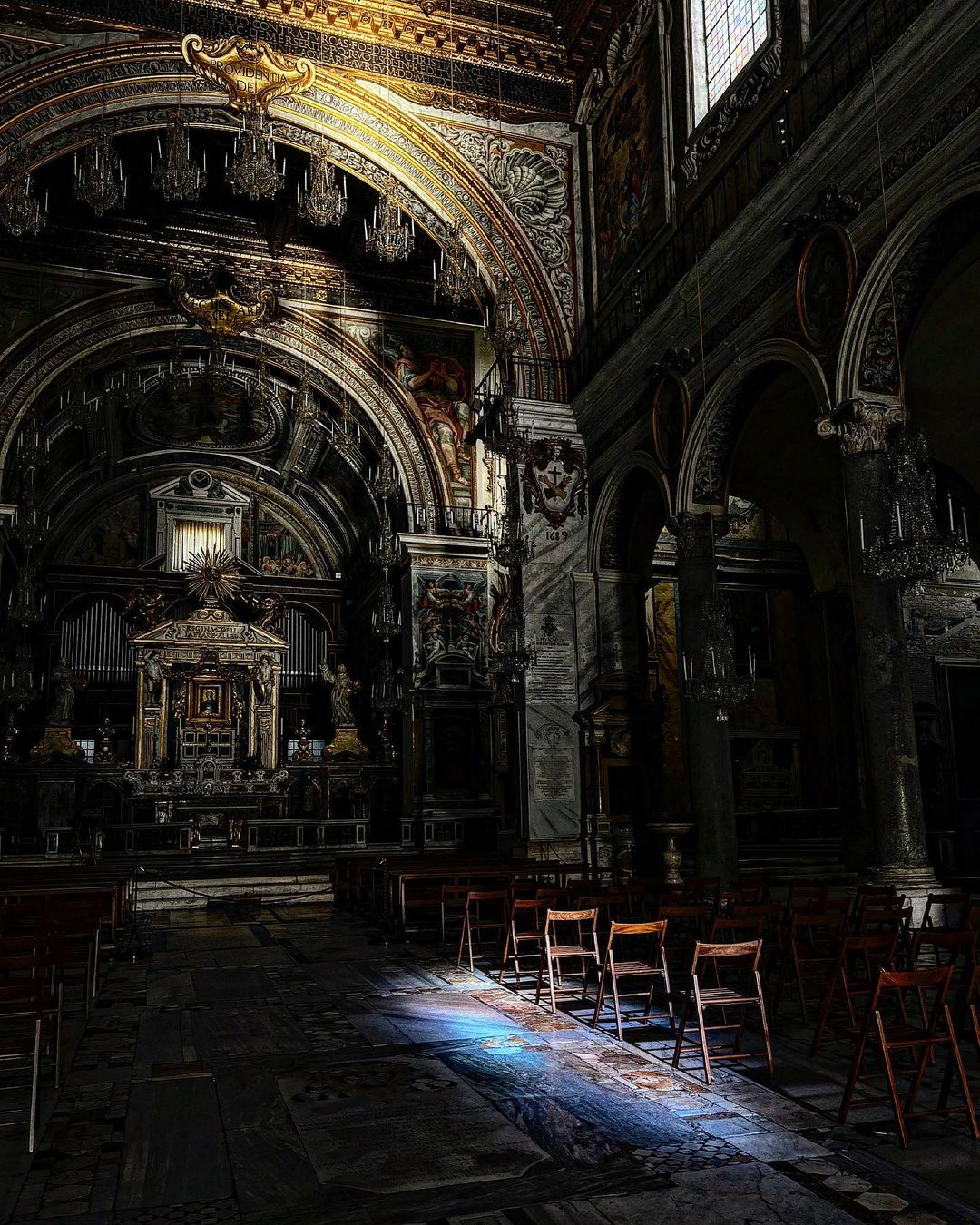 A striking interplay of light and shadow graces the interior of a historic church, where the dim ambience is punctuated by a shaft of sunlight streaming through a high window. The light caresses the church's intricate details, highlighting the golden ornate ceiling and the elaborate artwork that adorns the walls. Rows of humble wooden chairs face the altar, which stands as a silent testament to centuries of devotion. The gleaming marble floor, etched with the wear of time, reflects the solitary patch of blue light, adding a touch of ethereal beauty to the solemn atmosphere of this sacred space.