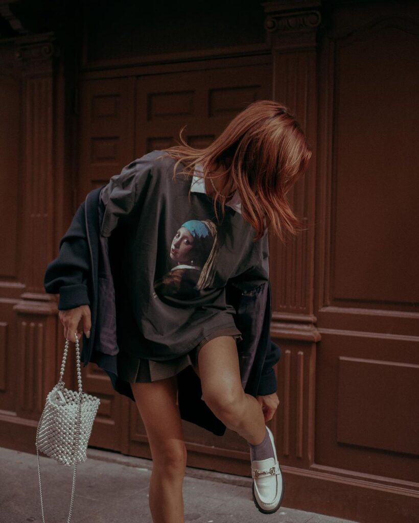 A dynamic street-style moment is captured in Madrid, Spain, as a woman strides confidently across the frame. Her movement is accentuated by her flowing auburn hair and the billow of her oversized, graphic-print sweatshirt featuring a classic art piece. She sports a matching miniskirt and complements her outfit with a chic, pearlescent beaded bag and sleek, silver loafers. The image is a blend of contemporary fashion and the timeless allure of Madrid's architectural charm, representing the vibrant and artistic spirit of the city.
