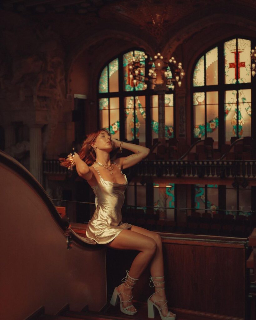 In the hallowed ambiance of a Barcelona church, a woman exudes a blend of reverence and modern style. She is seated on a wooden pew, the warm glow of the setting sun illuminating her silk dress and white strapped heels. Her pose is one of contemplation, her gaze lifted heavenward, hand gently resting on a bouquet of white flowers. The church's ornate architecture, with its intricate vaulted ceilings and vibrant stained glass windows, frames her elegantly. This setting creates a striking juxtaposition between the historic spirituality of the space and the contemporary fashion she embodies. The photograph captures a moment of stillness and the beauty of contrast within the sanctuary's ancient walls.