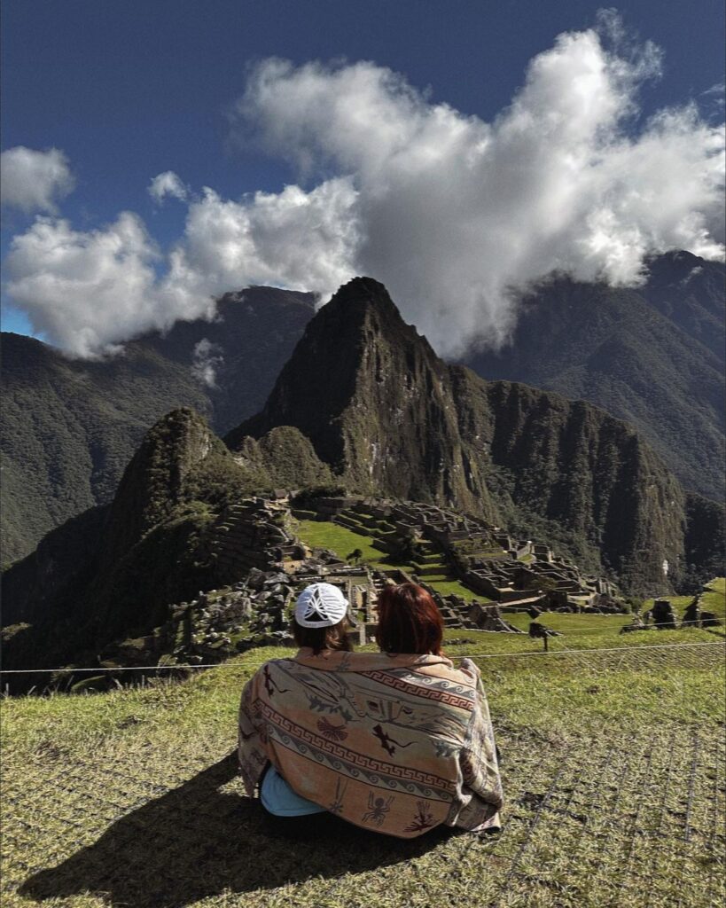 A serene and touching scene overlooking the ancient Inca city of Machu Picchu in Peru. A pair, identified as mother and daughter, sit closely together, sharing a moment of quiet contemplation. They are wrapped in a traditional Andean blanket, rich in cultural patterns, symbolizing a connection to the land and its history. The majestic Machu Picchu stands timeless before them, shrouded partially by the morning mist, while above, the clouds part to reveal a brilliant blue sky. This powerful image captures the essence of discovery and the intimate bond shared in the presence of one of the world's wonders.