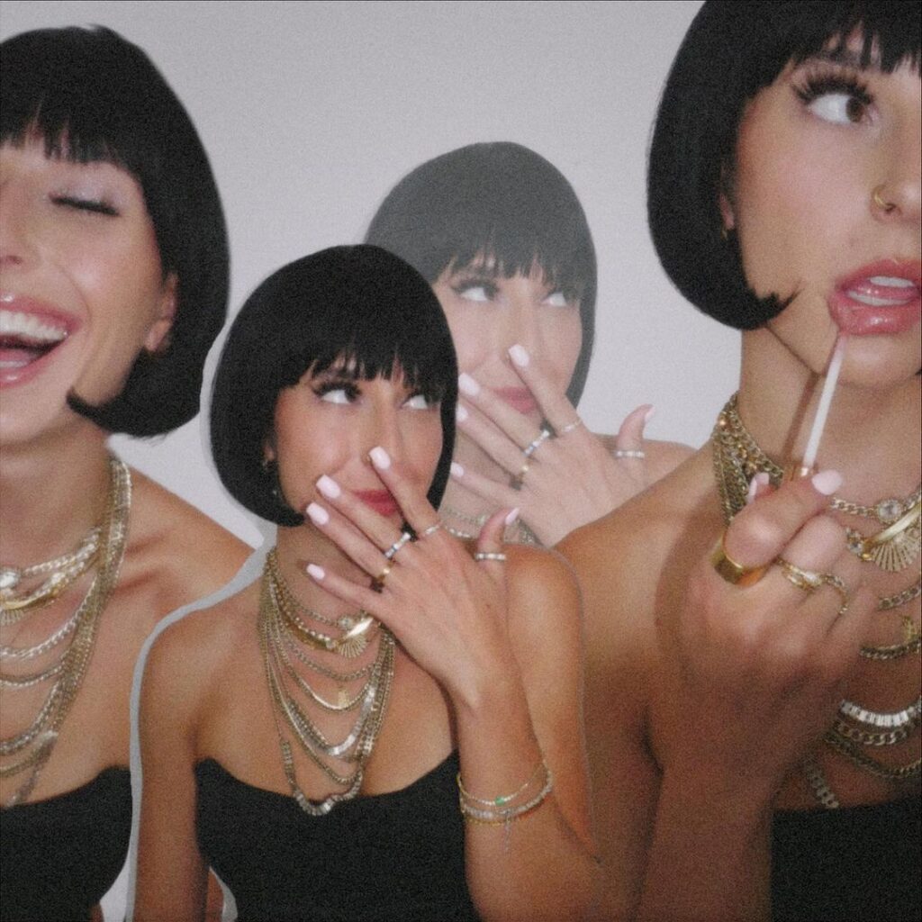 A creative and playful photo features a woman with a chic bob haircut in three overlapping poses, capturing laughter, a coy smile, and a moment of contemplation. Her style is accented by an array of gold necklaces and bracelets, and her nails are painted in a complementary shade. The lollipop in her hand adds a whimsical touch to the image. The layered composition creates a sense of movement and energy, showcasing her vibrant personality and fashionable ensemble against a plain white background.