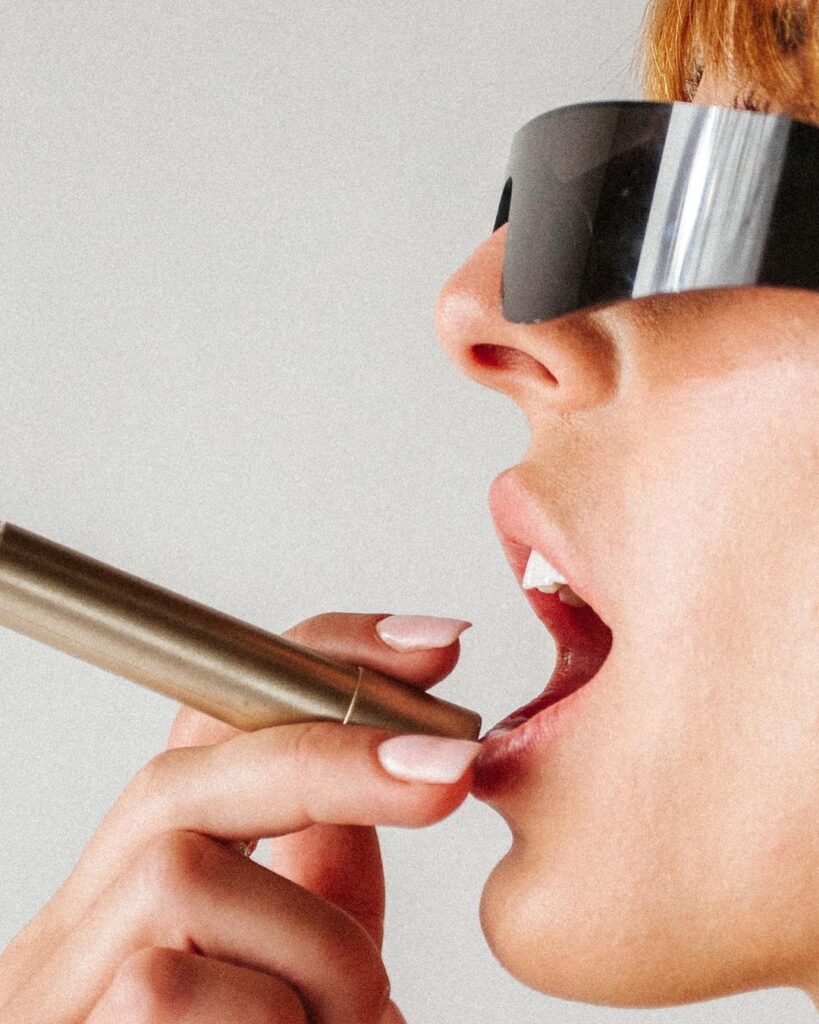 A close-up shot captures a modern, almost futuristic scene of a person applying cosmetics. They wear oversized, sleek, black visor sunglasses that exude a sense of contemporary fashion and mystery. The focus is on the precise application of lipstick, with a metallic gold tube held by a hand with neatly manicured, pale pink nails. The image is set against a neutral background, ensuring all attention is drawn to the action and style at the forefront. The photograph mixes elements of beauty routine with a hint of avant-garde, emphasizing detail and modernity.