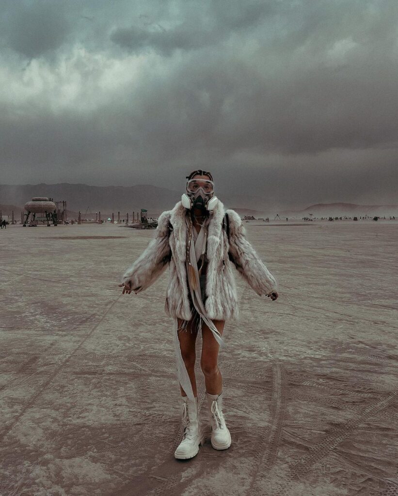 In the stark and ethereal landscape of Black Rock City at Burning Man, an individual stands in desert-appropriate garb that merges survival with expressionism. They are outfitted in a faux fur coat paired with a gas mask, prepared for the unpredictable dust storms of the Playa. The ensemble is completed with shimmering shorts and high white boots, striking against the muted tones of the desert. The ominous clouds above hint at the challenging conditions faced at the festival, yet there is an air of defiance and strength in the stance. This image captures the spirit of Burning Man: resilience, radical self-expression, and the unique community that arises in this temporary city.