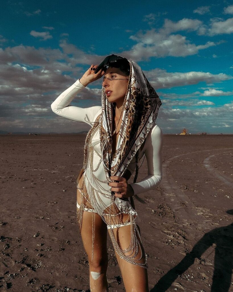 A woman in futuristic desert festival attire, standing on the dry playa at Burning Man, looks off into the distance. She wears a decorative white bodysuit adorned with chains and a patterned headscarf, holding aviator sunglasses to her forehead. The vast open landscape and scattered clouds convey a sense of adventure and exploration at the iconic event.